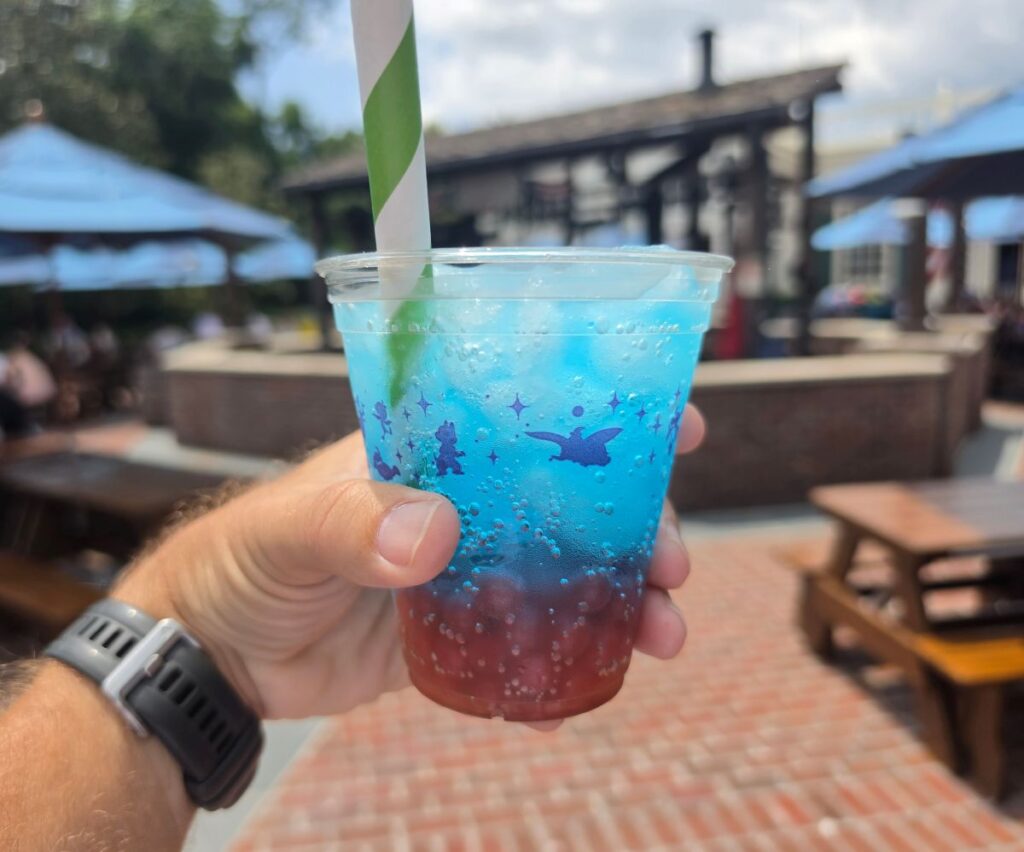 A hand holding a vibrant, layered red and blue iced beverage with a green striped straw, representing refreshing Fourth of July Treats served at an outdoor pavilion.