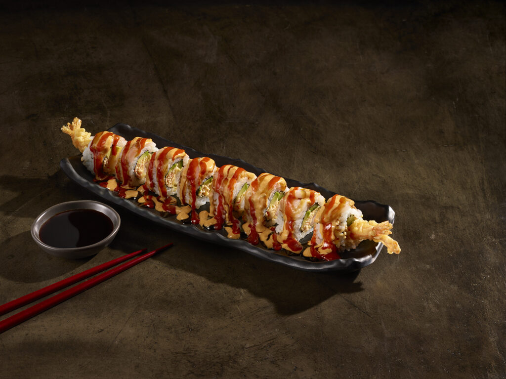A Tiger Roll sushi presentation on a long black wavy dish, showcasing shrimp tempura rolls heavily drizzled with spicy mayo and unagi sauce, served alongside a small dish of soy sauce and red chopsticks.
