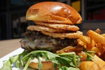 A gourmet truffle mushroom burger stacked with melted cheese, fresh arugula, caramelized onions, and crispy fried onion rings, served with a side of french fries from the city works chef selects menu.