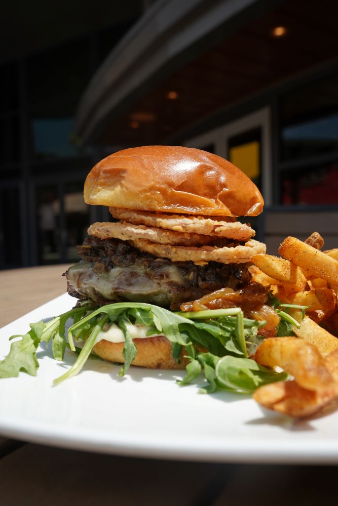 A gourmet truffle mushroom burger stacked with melted cheese, fresh arugula, caramelized onions, and crispy fried onion rings, served with a side of french fries from the city works chef selects menu.