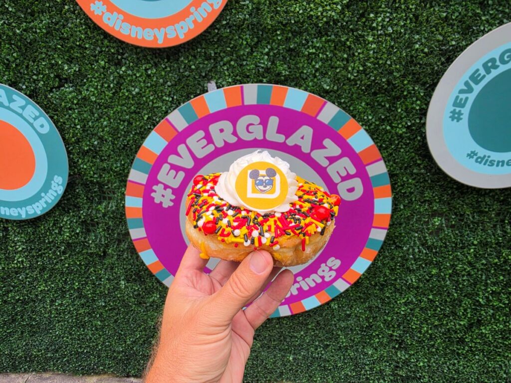 Hand holding a sprinkled donut with white frosting, atop a colorful plate that reads #EVERGLAZED, on green turf with other sign boards in the background.
