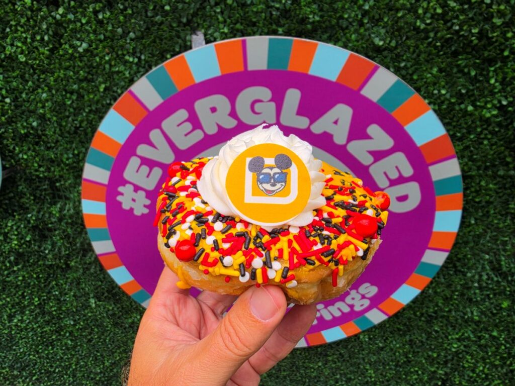 Donut with white frosting rosette and multicolor sprinkles held in a hand in front of a bright Everglazed sign plate with a hashtag theme.