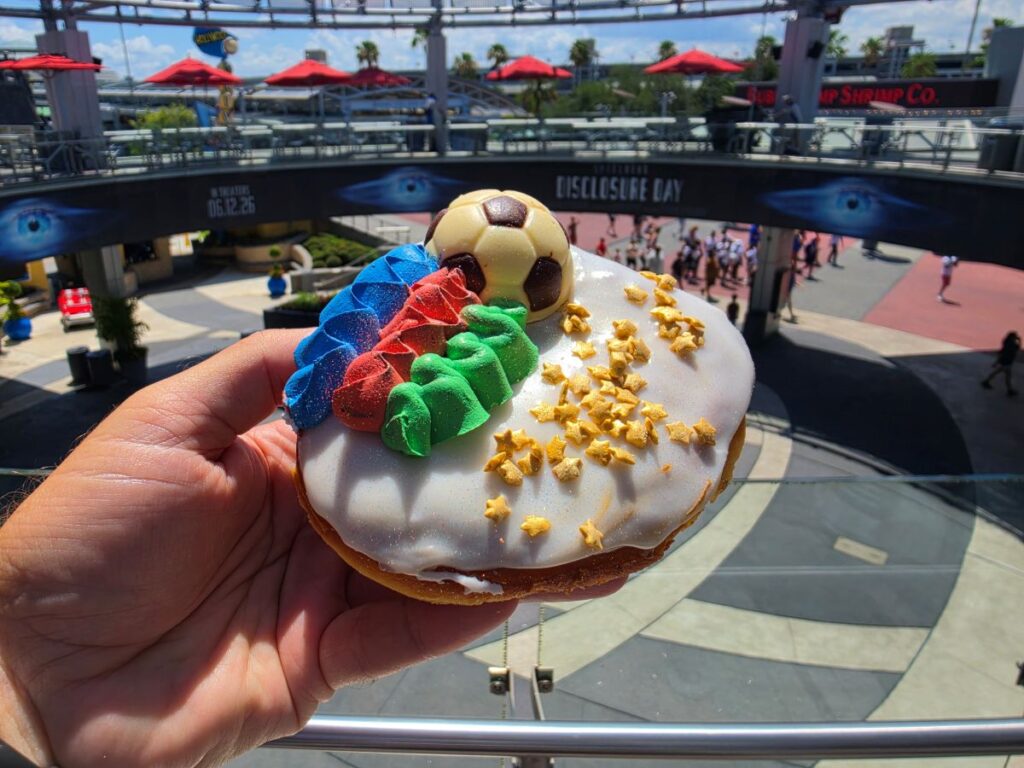 A person holding a World Cup Voodoo Doughnut at Universal CityWalk Orlando, with the open-air plaza and a "Disclosure Day" movie banner visible in the background. The large yeast doughnut features white icing, a white chocolate soccer ball, textured stripes of blue, red, and green frosting, and a dusting of edible gold star sprinkles and glitter.