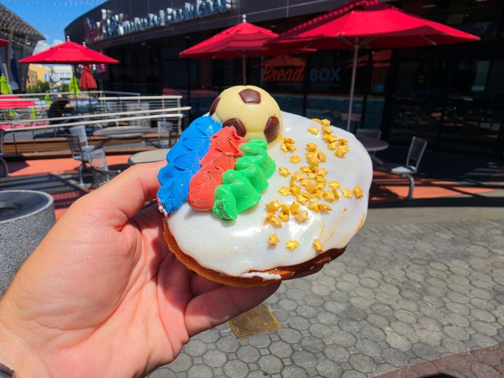 A close-up shot of a person holding a World Cup Voodoo Doughnut outside on a sunny day, with the red umbrellas and exterior of Bread Box Handcrafted Sandwiches in the background. The doughnut is topped with smooth white glazing, a detailed chocolate soccer ball, vibrant blue, red, and green frosting ridges, and scattered gold star sprinkle
