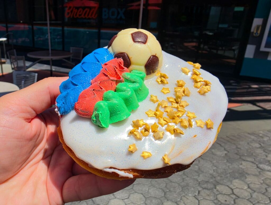 A hand holding a World Cup Voodoo Doughnut at an angle to showcase its thick, golden-brown fried dough base. The top is decorated with a white chocolate soccer ball, thick piping of blue, red, and green frosting, white icing, and shimmering gold stars under bright daylight.