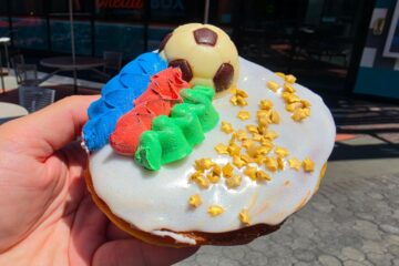 A hand holding a World Cup Voodoo Doughnut at an angle to showcase its thick, golden-brown fried dough base. The top is decorated with a white chocolate soccer ball, thick piping of blue, red, and green frosting, white icing, and shimmering gold stars under bright daylight.