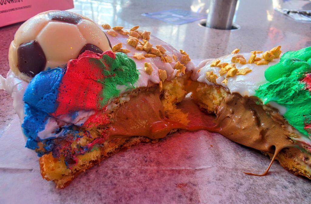 A World Cup Voodoo Doughnut sliced completely in half on a wax paper surface, revealing a rich, creamy chocolate cream filling oozing from the center. The vibrant blue, red, and green frosting, white icing, and white chocolate soccer ball remain visible on top of the cut pastry.
