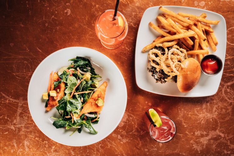 An overhead, flat-lay shot of a restaurant meal from the City Works Chef Selects menu, arranged on a textured copper tabletop. On the left, a white bowl holds a crisp salad featuring spinach, shredded carrots, and diced avocado atop triangular, golden-brown crispy rice cakes. On the right, a white plate presents a gourmet burger topped with melted cheese, mushrooms, and fried onion rings, served with a large side of french fries and a small cup of ketchup. Two colorful, iced cocktails with fruit garnishes—one orange-pink and one deep red—are positioned near the plates.