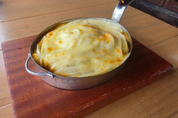 Cottage Pie shown in an oval metal pan with a handle. The top consists of creamy-looking potatoes for a Rose & Crown Review