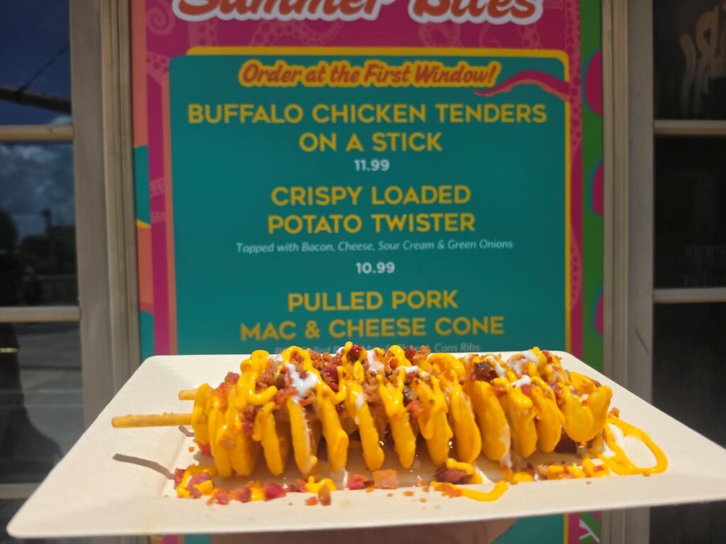 A close-up shot of a loaded potato twister on a paper plate held up in front of a colorful teal and pink "Summer Bites" menu board at SeaWorld Orlando. The menu lists item prices, including the "Crispy Loaded Potato Twister" for 10.99, establishing a direct connection between the food and its ordering location.