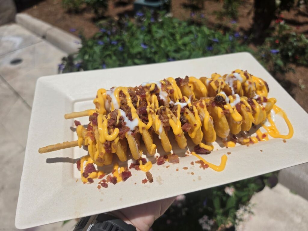 A close-up, high-angle shot of the Crispy Loaded Potato Twister spiral-cut potato onon two wooden skewers, served on a white rectangular paper plate. The crispy, golden-brown potato spiral is heavily drizzled with bright yellow cheese sauce and white sour cream, then topped with a generous scattering of real bacon bits at SeaWorld Orlando.