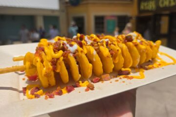 A top-down, slightly angled view of a crispy spiral potato twister served on a white paper tray. The food is generously drizzled with warm cheese sauce and white cream, packed with bacon bits, with an outdoor garden area featuring green foliage and purple flowers visible in the background.