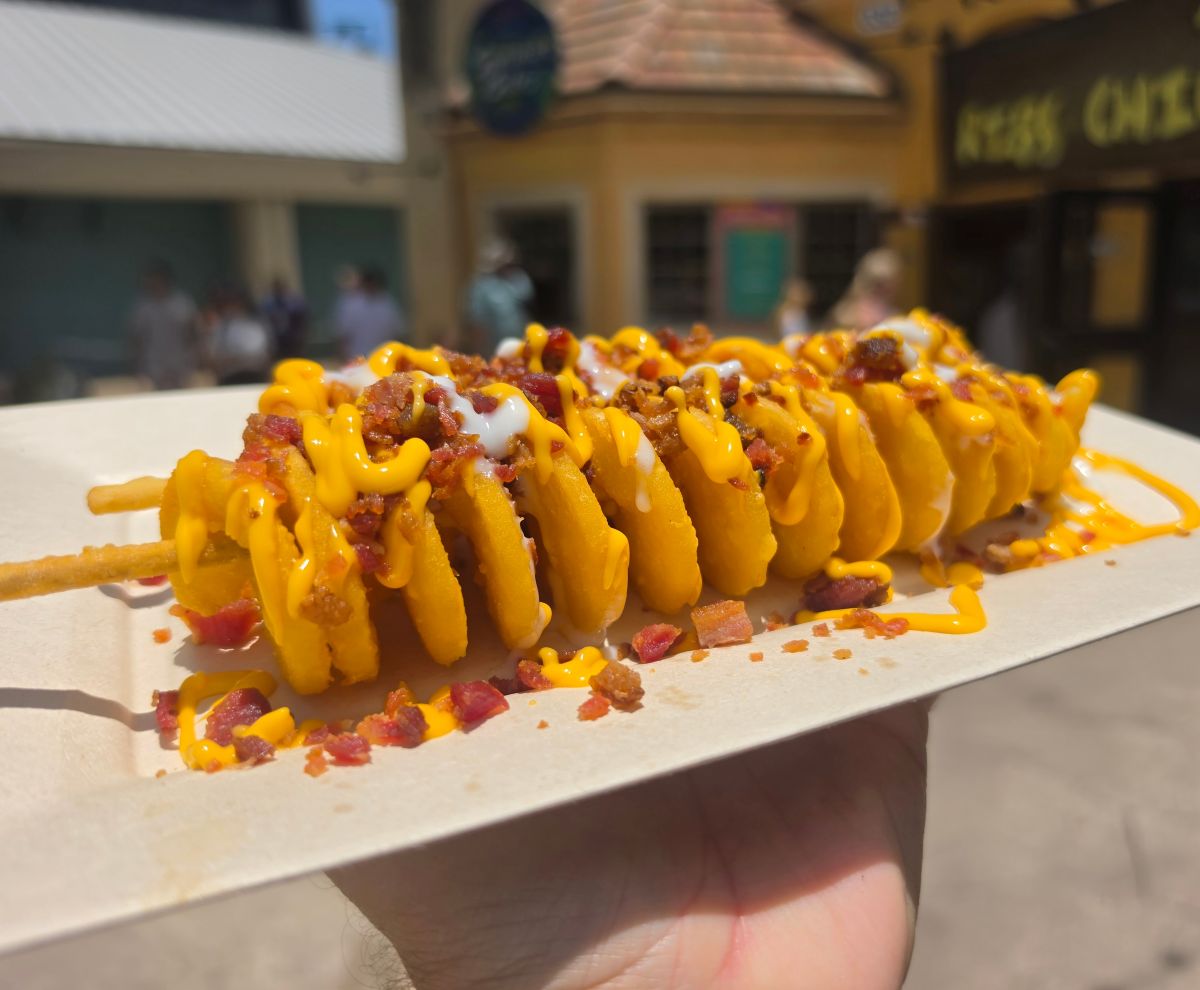 A top-down, slightly angled view of a crispy spiral potato twister served on a white paper tray. The food is generously drizzled with warm cheese sauce and white cream, packed with bacon bits, with an outdoor garden area featuring green foliage and purple flowers visible in the background.