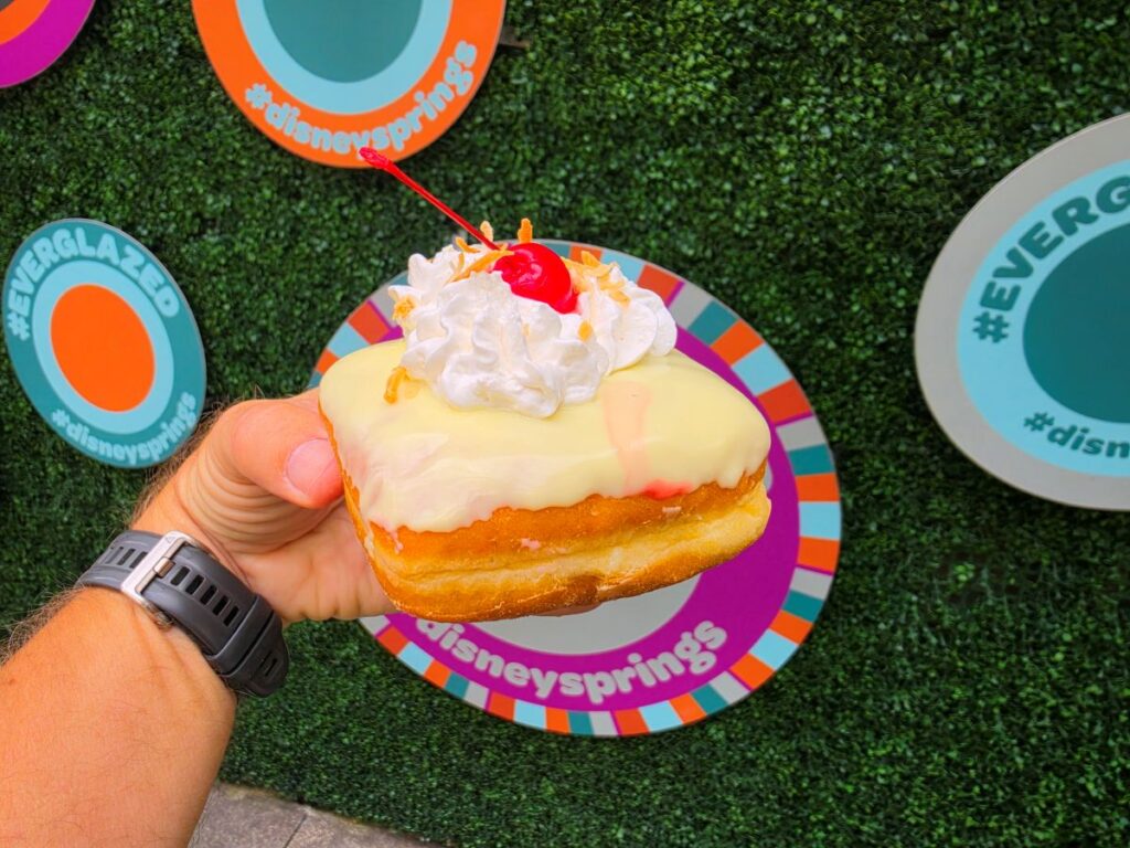 Hand holding a glazed donut topped with whipped cream, a cherry, and crunch toppings over a bright striped plate with Disney Springs branding in the background for Everglazed Disney Springs review