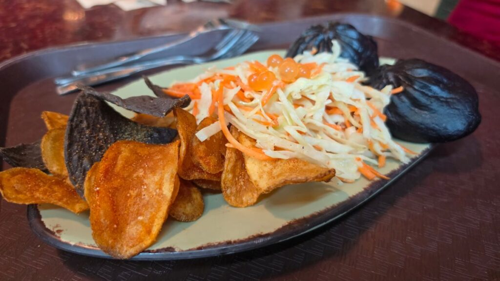 A restaurant tray holding a plate with two dark-colored spicy pork pods, a side of fresh vegetable slaw garnished with orange pearls, and a colorful mix of seasoned potato and tortilla chips.