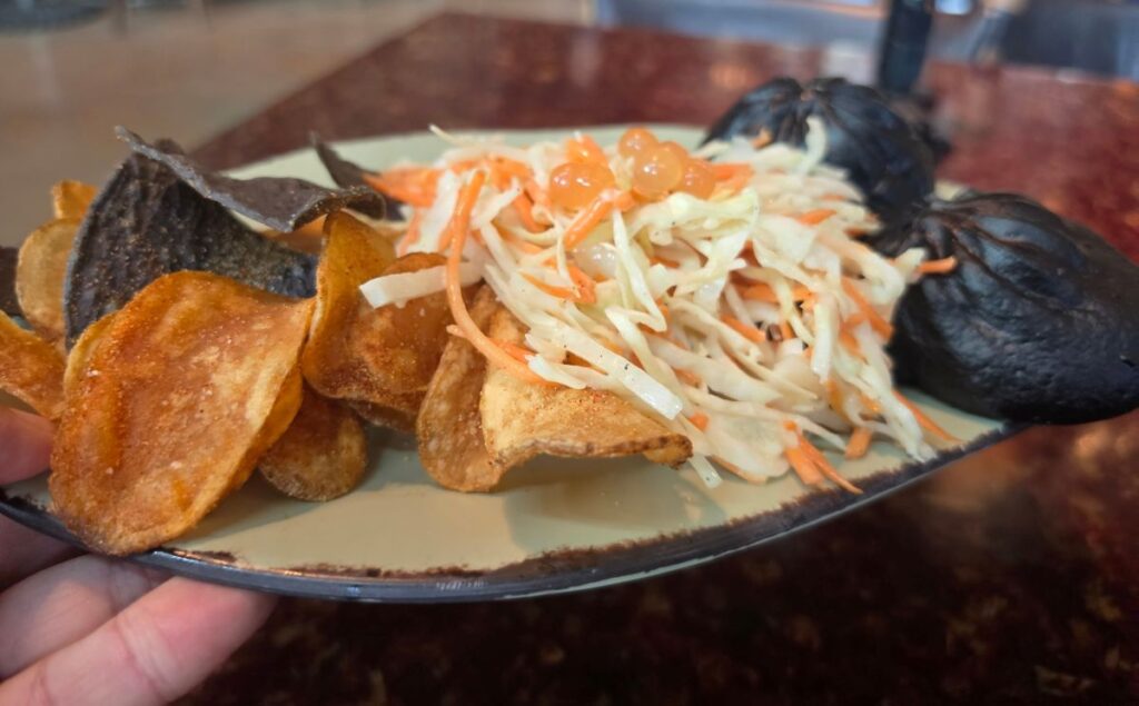 A hand holding a plate featuring two dark purple spicy pork pods paired with a crisp cabbage and carrot slaw topped with orange popping boba pearls, alongside a side of crunchy seasoned potato chips.