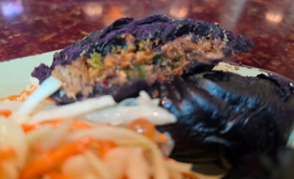 A close-up, cross-section view of one of the sliced spicy pork pods, showing a hearty filling of seasoned shredded pork and green vegetables inside a vibrant, dark purple steamed bun.
