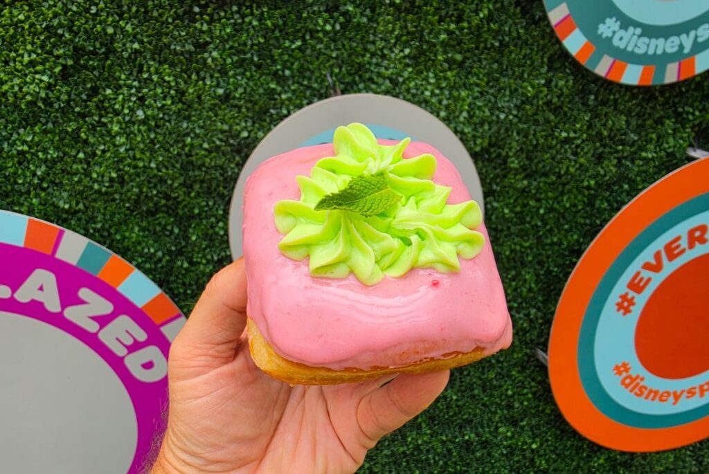 Hand holding a pink frosted donut topped with lime-green icing swirls and a mint leaf, against a colorful background.