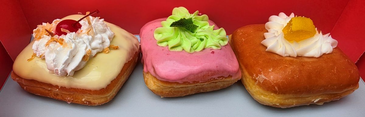 Three frosted square donuts in a red box: left with white glaze, whipped cream, and cherry; middle pink glaze with green rosette; right orange glaze with whipped cream and candied orange for an Everglazed Disney Springs review