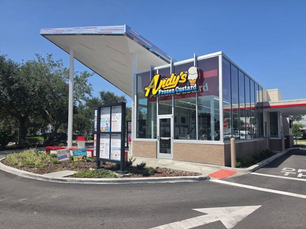 Exterior view of the modern, glass-walled Andy's Frozen Custard building with drive-thru menu boards and a large retro roof canopy.