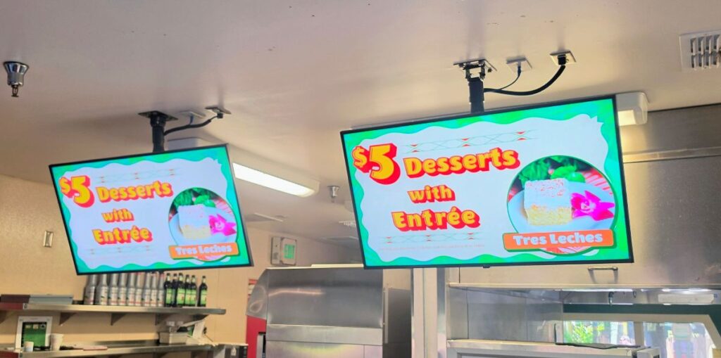 Two overhead digital menu screens inside a theme park restaurant displaying a five dollar desserts with entree promotion highlighting Tres Leches cake next to an image of the dessert.