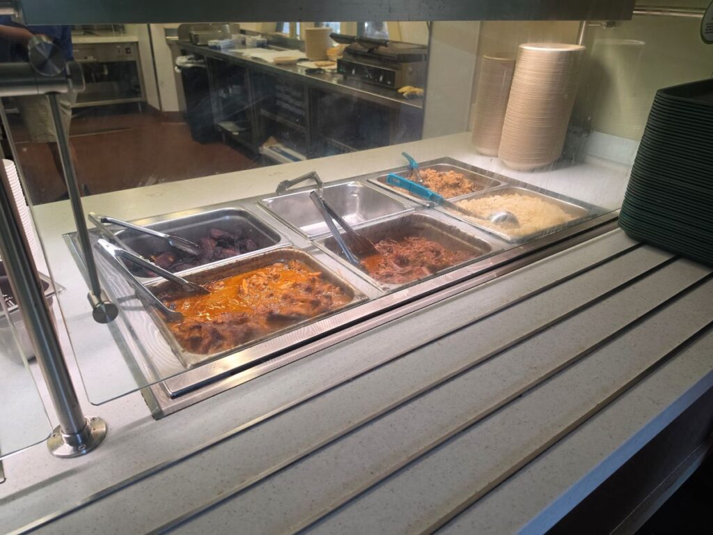 A theme park cafeteria service counter behind a glass shield featuring stainless steel trays filled with traditional Cuban food, including shredded beef, yellow rice, and plantains.