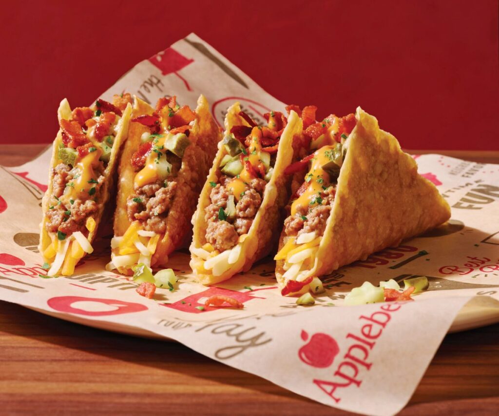 Four Applebee's tacos lined up on branded parchment paper, filled with ground meat, shredded cheese, melted cheese sauce, bacon bits, and diced pickles for Applebee's 2 for $25