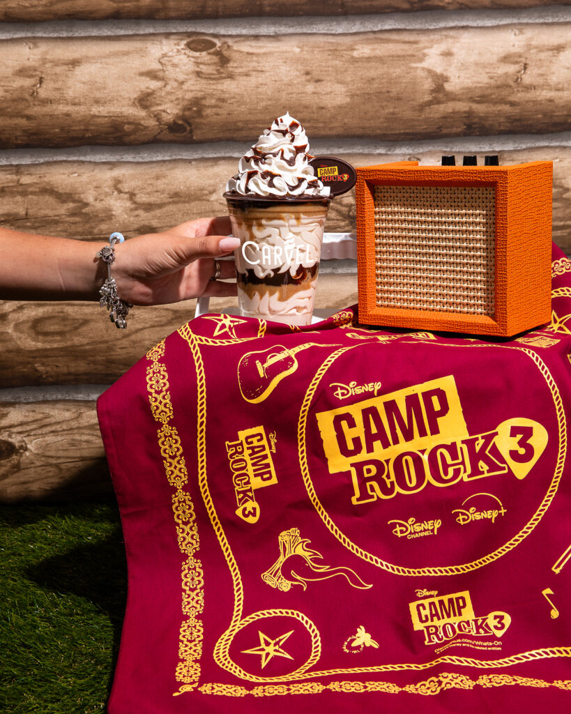 A hand holds a Carvel sundae next to an orange portable speaker on a red Camp Rock 3 bandana, all set against a rustic log wall and green grass.