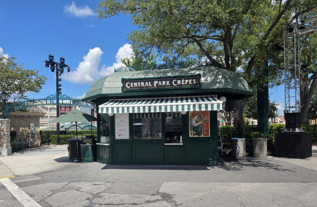 A green quick-service food kiosk named Central Park Crêpes at Universal Studios Florida, with a green-and-white striped awning, displaying a menu and set against a backdrop of trees and a bright blue sky.