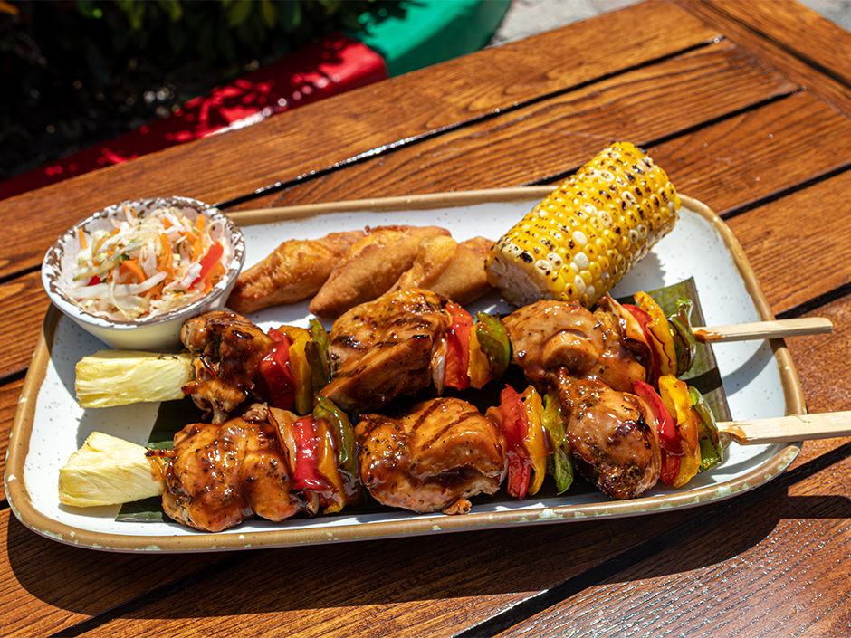 Grilled chicken skewers served with peppers, pineapple, roasted corn on the cob, festival bread, and coleslaw