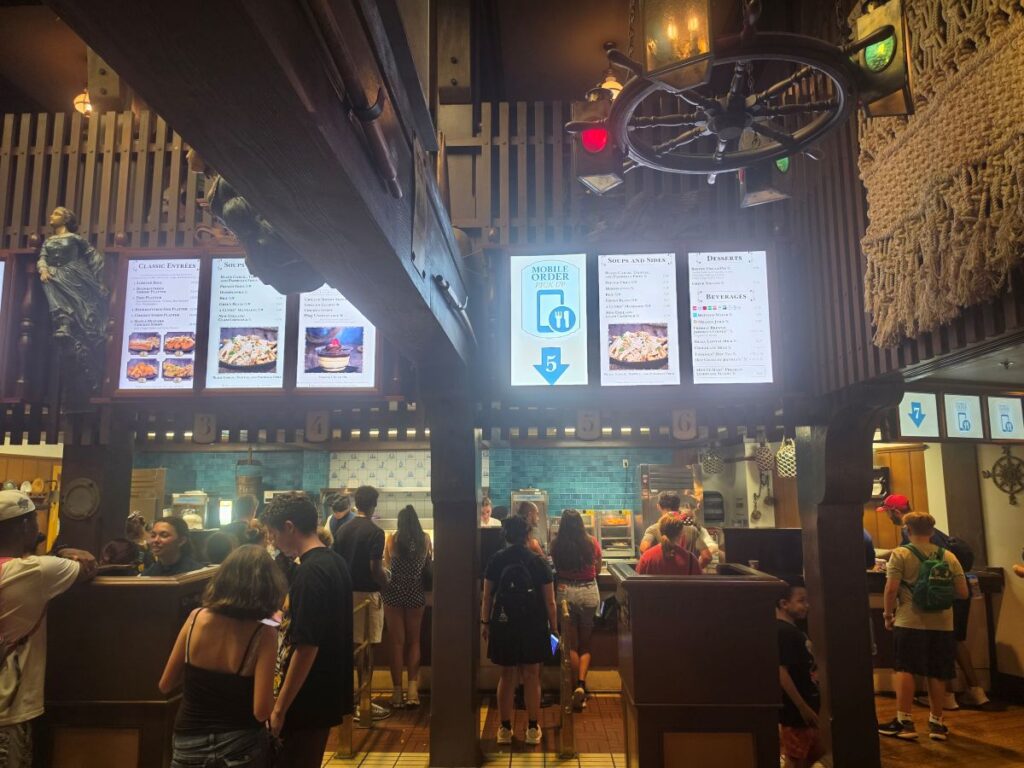 Interior view of the ordering counter and Mobile Order pick-up area inside Columbia Harbour House at Magic Kingdom, featuring nautical décor and digital menu boards.