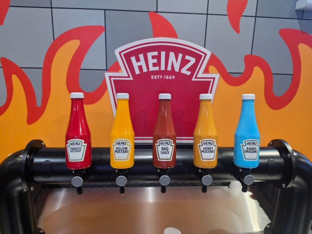 A Heinz condiment dispenser station featuring five upside-down squeeze bottles mounted on a black dispenser pipe against an orange flame background. From left to right, the bottles are labeled Tomato Ketchup, Yellow Mustard, BBQ Sauce, Honey Mustard, and blue-colored Ranch Dressing.