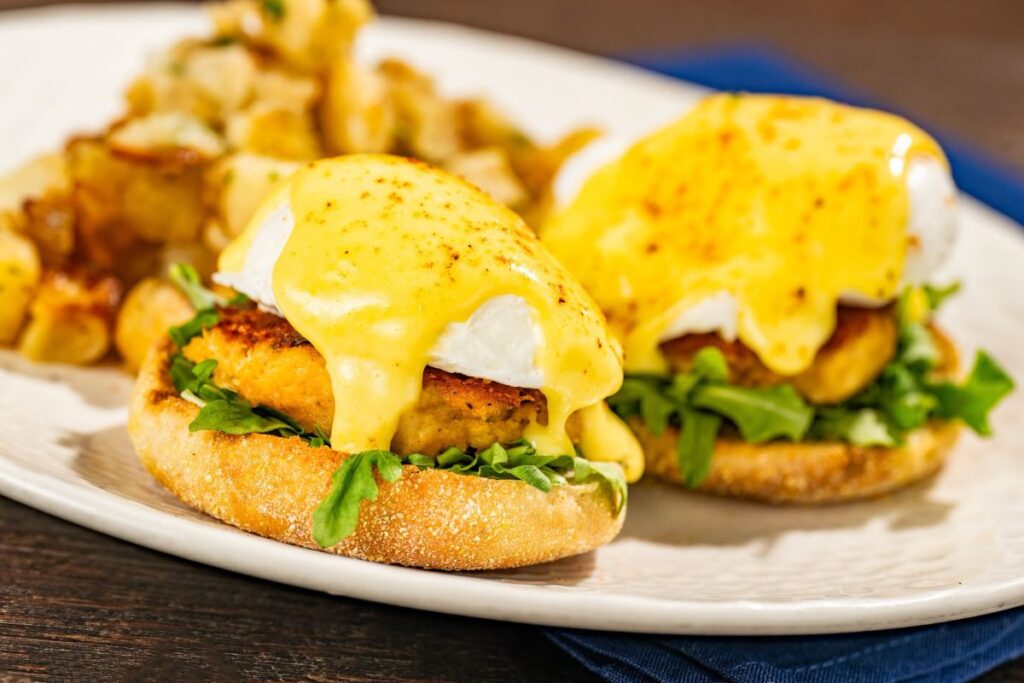 Crab Cake Eggs Benedict with poached eggs and hollandaise sauce served with side potatoes at Ale & Compass breakfast.