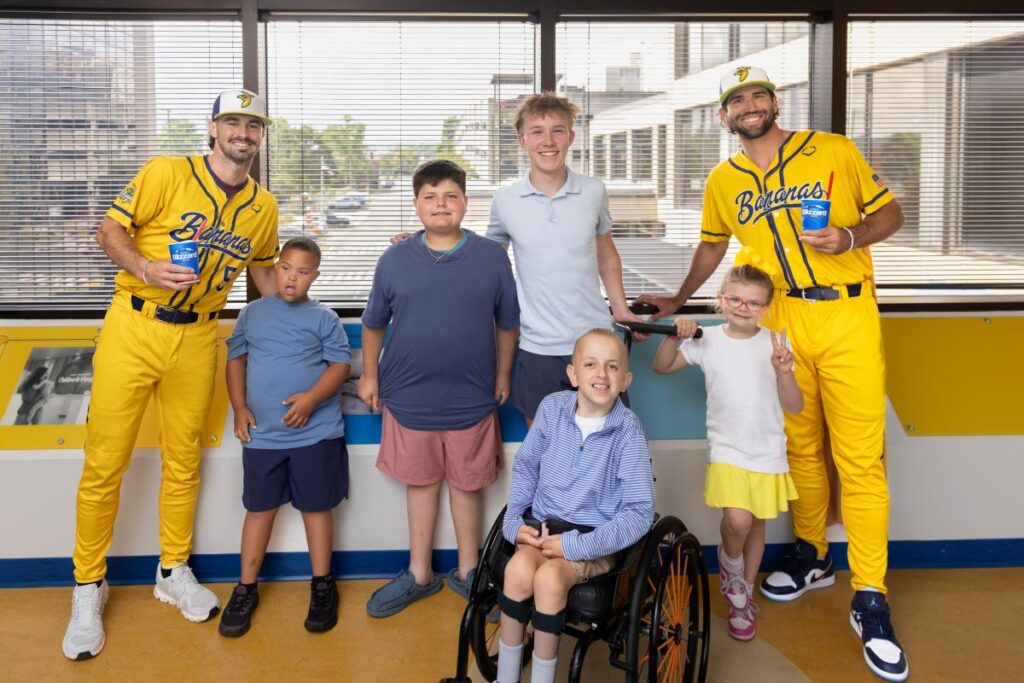 Two baseball players in yellow Bananas uniforms stand with five children, one in a wheelchair, posing for a photo in front of a windowed wall.