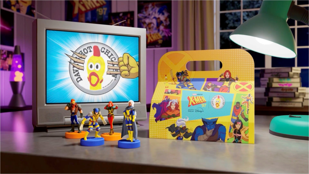 A retro-themed product shot showcasing the Dave's Hot Chicken X-Men '97 promotion on a dark desk. On the left, an old-school silver CRT television screen displays the Dave's Hot Chicken rubber chicken logo being slashed by Wolverine's claws. Four small X-Men collectible toy figures are lined up on the desk in front of the TV. To the right sits the yellow, illustrated X-Men meal box under the glow of a green desk lamp. In the blurry background, a purple lava lamp glows on the left, and a stack of VHS tapes rests on the right.