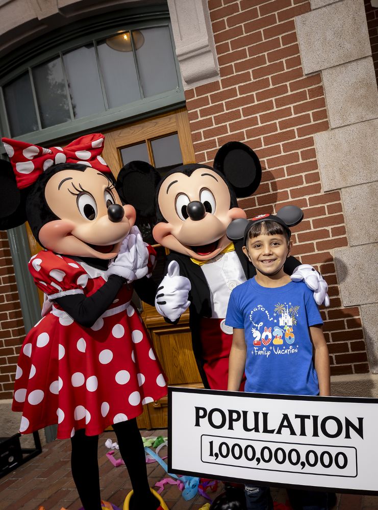 A young boy wearing Mickey Mouse ears and a blue Disney family vacation t-shirt smiles widely as he stands between Minnie Mouse and Mickey Mouse. Minnie, in her red and white polka dot dress, beams with her hands to her face, while Mickey gives a thumbs-up. In front of them, the boy holds a white sign that reads "POPULATION 1,000,000,000" against a brick building backdrop.