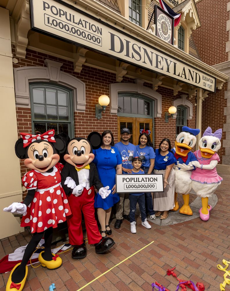 A wide shot in front of the Main Street Disneyland Railroad station showing a family of four posing with Disney characters to celebrate the 1 billionth guest milestone. Minnie Mouse, Mickey Mouse, Donald Duck, and Daisy Duck stand alongside two Disney Cast Members and the family. The young boy holds a sign that reads "POPULATION 1,000,000,000" while standing under a large station sign displaying the same 1 billion population milestone.