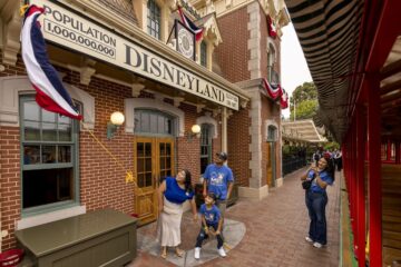A family of three stands outside the brick Main Street Disneyland Railroad station next to a red train car. A woman and a young boy playfully pull on a yellow rope connected to a flying flag, while a man next to them looks up and smiles. A few feet away, another woman captures the moment on her smartphone. The station marquee above them displays a sign reading "POPULATION 1,000,000,000".
