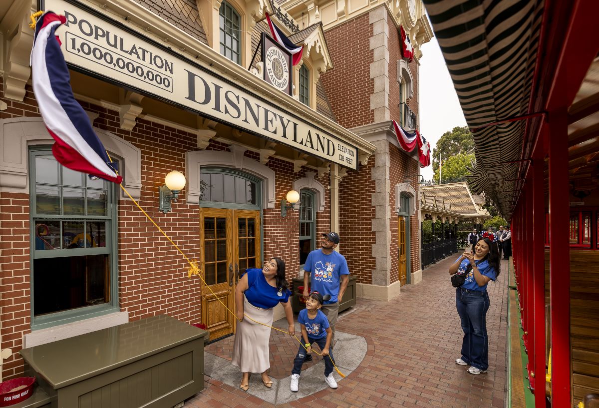 A family of three stands outside the brick Main Street Disneyland Railroad station next to a red train car. A woman and a young boy playfully pull on a yellow rope connected to a flying flag, while a man next to them looks up and smiles. A few feet away, another woman captures the moment on her smartphone. The station marquee above them displays a sign reading "POPULATION 1,000,000,000".