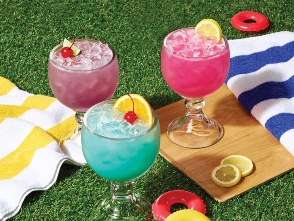 Three colorful summer cocktails—purple, blue, and pink—served in large goblet glasses with ice and fruit garnishes on a grassy picnic setup.