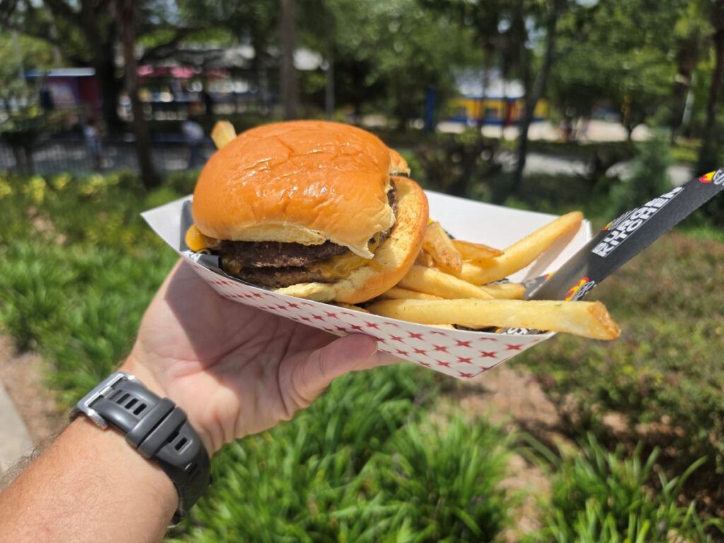 A close-up view of a hand holding a red-and-white patterned paper tray filled with a double cheeseburger and golden french fries, with a black Burger Kitchen wrapper tucked underneath.