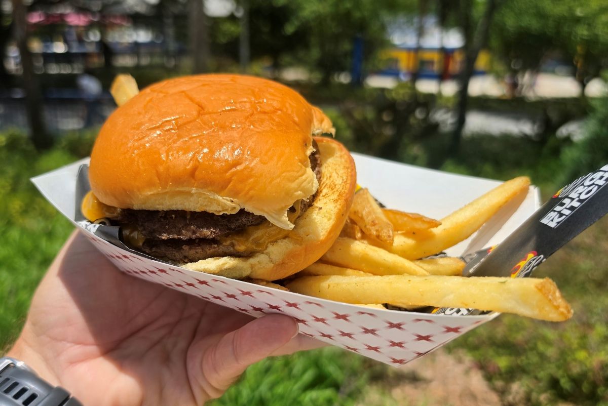 A close-up shot of a double cheeseburger with melted yellow cheese and a side of french fries being held in a checkered paper boat outdoors.