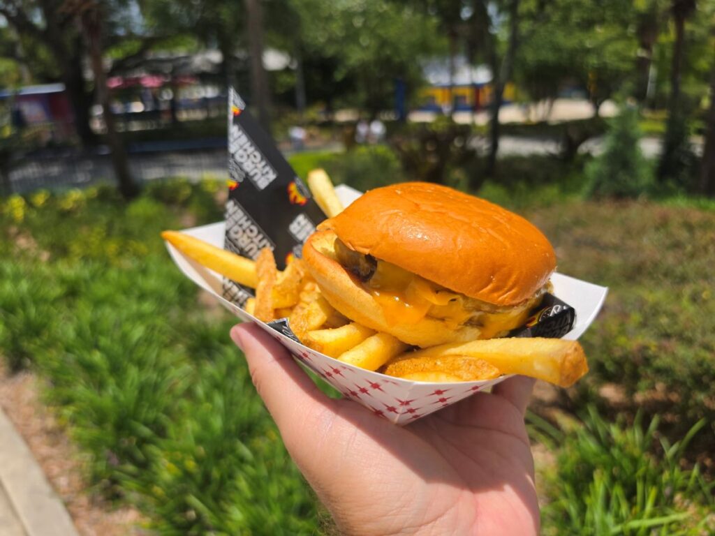 A hand holding a paper food boat containing a double cheeseburger on a soft bun and a side of french fries. The background shows a blurry outdoor theme park setting with green trees and lawns.