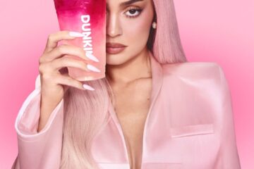 Kylie Jenner with pastel pink hair and a light pink blazer holding a Dunkin' pink iced beverage over one eye against a solid pink backdrop.