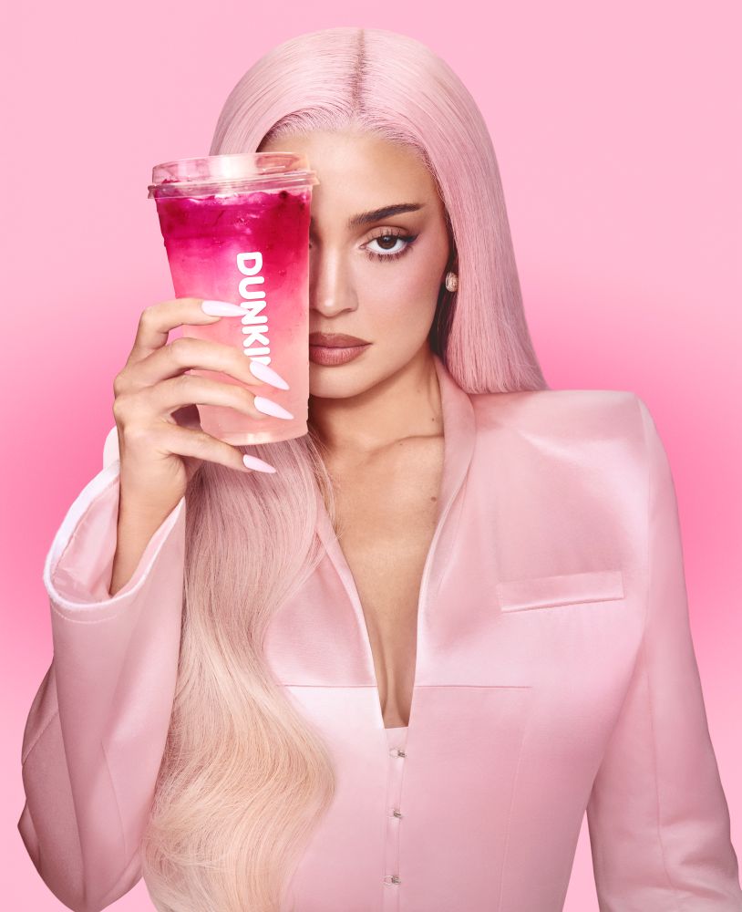 Kylie Jenner with pastel pink hair and a light pink blazer holding a Dunkin' pink iced beverage over one eye against a solid pink backdrop.