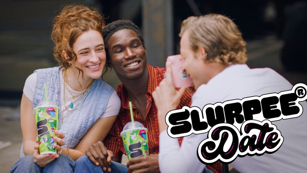 Three friends celebrating 7-Eleven Free Slurpee Day. A woman with red hair and a man in a red button-down shirt smile closely together, holding vibrant green Slurpee cups with lime green straws, while a third person captures the moment with a pink camera for Free Slurpee Day 2026rFree