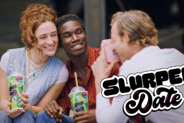 Three friends celebrating 7-Eleven Free Slurpee Day. A woman with red hair and a man in a red button-down shirt smile closely together, holding vibrant green Slurpee cups with lime green straws, while a third person captures the moment with a pink camera.