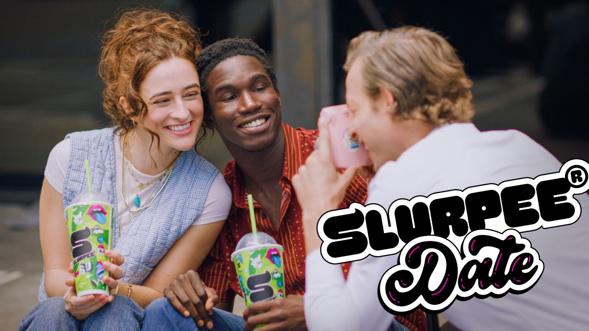 Three friends celebrating 7-Eleven Free Slurpee Day. A woman with red hair and a man in a red button-down shirt smile closely together, holding vibrant green Slurpee cups with lime green straws, while a third person captures the moment with a pink camera.