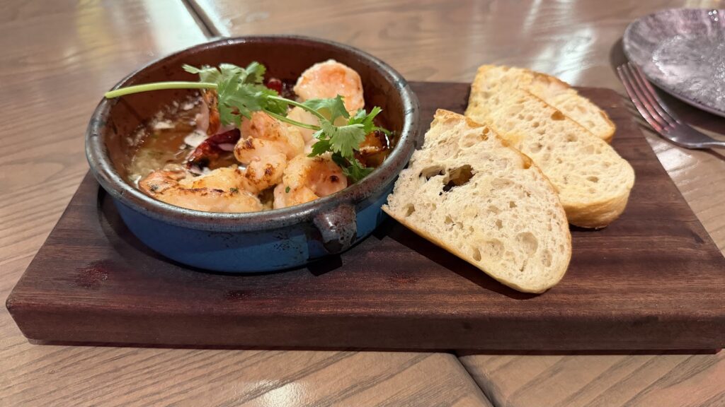 Close-up of Gambas al Ajillo, featuring sautéed shrimp garnished with fresh cilantro, in a blue ceramic dish, served next to sliced, crusty bread on a wooden board for a Toledo Disney review.