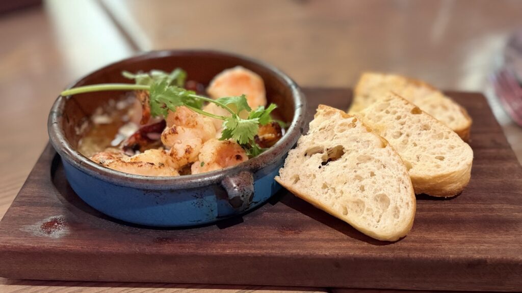 Sautéed shrimp with cilantro in a blue dish alongside three slices of bread on a wooden serving board, captured from a slightly wider angle.